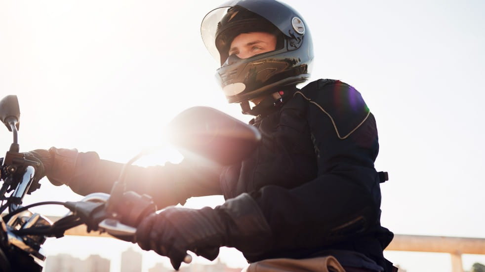 Male on a motorbike, wearing helmet, visor is in upward position, both hands on the handlebars. Sunlight streams over his arms and across handlebars. 